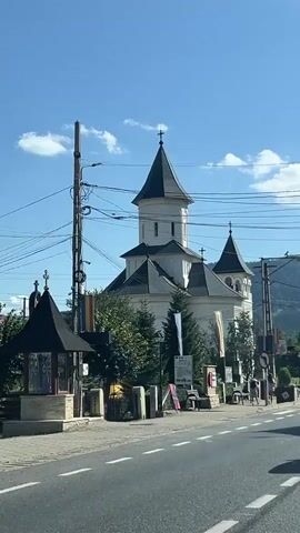The video shows a white church with dark spires under a clear blue sky. There are trees, utility poles, and power lines in front of the church. A road with white lines is in the foreground. Several people are walking on a sidewalk in front of the church. There are also benches and flower pots. In the background, there are hills covered with trees.