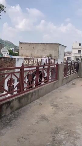 The video pans across a rooftop scene. In the foreground, a concrete ledge with a reddish-brown metal railing runs horizontally. Beyond the railing, there are buildings. To the left, a white building with a satellite dish is visible. Further back, a two-story building with a flat roof and several windows is present. To the right, a brick building with a water tank on top is shown. The ground of the rooftop is concrete, with a few scattered bricks and a coiled black hose. The sky is partly cloudy. There are no visible people or animals in the video.