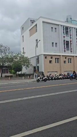 A street scene with a cloudy sky. On the left, a white and gray modern building with windows is visible. Across the street, there is a sidewalk lined with trees and parked motorcycles. A few motorcycles are in motion on the street, including a white scooter and a blue motorcycle carrying a rider wearing a helmet. To the right, another building with a prominent Starbucks logo is seen, also with a line of parked motorcycles in front. The street has yellow and white lane markings. The overall impression is an urban environment on an overcast day.