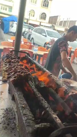 This video captures a close-up view of satay being grilled over an open flame. The satay skewers are arranged horizontally on a grill, with visible flames licking the meat. In the background, a person in a dark t-shirt with a colorful pattern is tending to the grill. Behind them, there are orange plastic chairs and tables, suggesting an outdoor food stall or restaurant setting. A white minivan and a blue tarp are also visible in the distance. The lighting appears to be natural, possibly late afternoon. The overall scene depicts a food preparation process with a focus on the cooking of the satay.