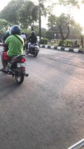 The video shows a street scene with several motorcycles moving in the same direction. The camera appears to be mounted on a moving vehicle, likely another motorcycle, as indicated by the motion blur. The foreground shows the road surface with some debris. In the middle ground, several motorcycles with riders are visible. One rider in the immediate foreground is wearing a green shirt and a blue helmet, and is moving away from the camera. Further ahead, other motorcycles are also in motion. The background consists of trees lining the road and a hazy sky, suggesting it might be early morning or late afternoon. The road appears to have multiple lanes, separated by white lines and a curb on the right side. The overall impression is of a common street scene with motorcycle traffic.