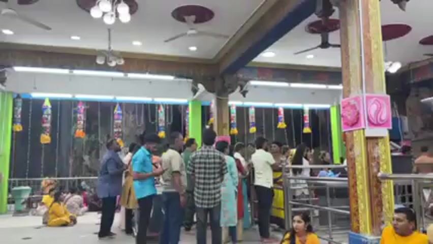The video shows the interior of a decorated temple or shrine. A group of people, dressed in casual attire, are milling around inside. Some are standing, others are seated on the floor. A shirtless man carrying a tray walks through the crowd. Decorations hang from the ceiling and walls, including colorful garlands and painted panels. The atmosphere appears calm and reverent.