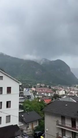 The video presents an overcast view of a European town nestled in a valley between mountains. The sky is heavy with gray clouds, and mist clings to the upper reaches of the densely forested mountains. Houses with varied roof colors are visible in the foreground and midground, and a few cars are parked on the streets. The overall atmosphere suggests a rainy or damp day.