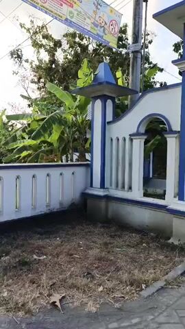 The video shows a cemetery entrance with a sign that reads "MAKAM UMUM TUNENGGUNG SURONTANI DS WAJAK KIDUL KEC. BOYOLANGU KAB. TULUNGAGUNG". The entrance has blue and white pillars and an arched gateway. A person wearing a black shirt and helmet is standing in front of the entrance, taking a photo with their phone. There are motorcycles parked to the left of the entrance. The environment appears to be a rural area with trees and a clear sky. There is also a banner with children's illustrations hanging from a utility pole. The ground in front of the entrance is covered with dry leaves and debris.
