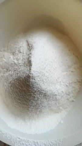 A white bowl sits on a speckled gray countertop. Inside the bowl is a pile of white powder, likely flour and sugar, with a distinct division creating two separate mounds. The lighting is bright, casting subtle shadows within the bowl. The camera is positioned directly above the bowl, offering a top-down view.