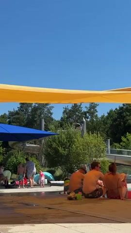 The video shows a group of people sitting on the edge of a pool on a sunny day. A large yellow canopy shades the area. In the background, there are trees, a water slide, and other pool structures. Several people are visible, including adults and children, some of whom are in swimsuits. One person appears to be holding a baby or a small child. There are toys and bags scattered around the edge of the pool. The sky is clear and blue.