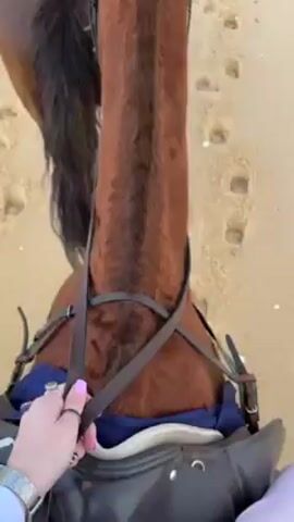 The video shows a group of people horseback riding on a sandy beach. The camera is mounted on one of the horses, providing a first-person view of the ride. The horse is brown, with a black mane and tail.  The rider's hands are visible, holding the reins. Other riders and horses can be seen ahead, riding along the shoreline. The ocean is to the side, with gentle waves rolling onto the beach. The sky is partly cloudy. The beach is covered with footprints from both humans and horses.