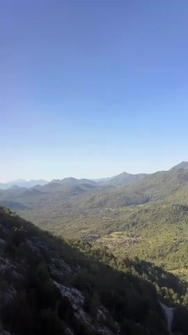A vertical video captures a scenic view of rolling green mountains under a clear blue sky. The camera pans slightly, revealing a valley dotted with small buildings and a winding road. The foreground is dominated by the steep, rocky slope of a mountain covered in dark green trees. Sunlight illuminates the landscape, creating shadows and highlighting the textures of the terrain. There are no people or animals visible in the video. The overall impression is one of a remote, natural landscape bathed in warm daylight.
