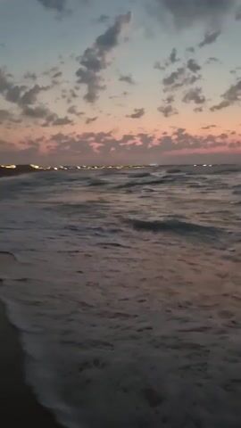 The video shows a body of water with waves gently lapping onto a sandy shore. The sky above is a dusky twilight with pink and orange hues, dotted with scattered clouds. In the distance, a line of lights suggests a populated area or cityscape. The overall impression is of a serene beach scene at dusk or dawn. There are no people or distinct objects visible in the immediate foreground or on the water.