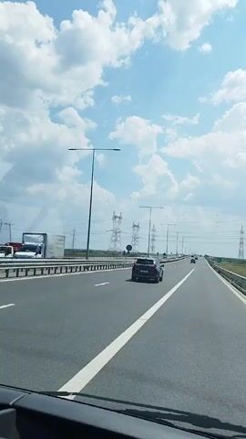 The video shows a view from inside a vehicle traveling on a highway. The road is paved and has white lane markings. Several cars are visible on the highway, moving in the same direction. On the right side of the road, there is a grassy area and a barrier. In the distance, there are numerous tall power line towers stretching across the landscape. The sky is blue with fluffy white clouds. The overall impression is of a typical highway journey on a clear day.