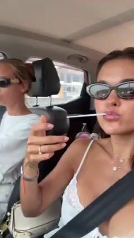 Two young women are in a car. The driver, with blonde hair pulled back in a bun, wears sunglasses and a white top. The passenger, with brown hair, also wears sunglasses and a white top with lace details. She has a white stick in her mouth, possibly a lollipop or straw, and is holding a white container. Both are wearing seatbelts. The car's interior is visible, including the dashboard and seats. The background shows an urban environment with buildings. The passenger is holding the camera and seems to be recording a selfie video. The driver appears focused on the road.