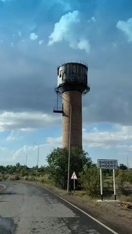 The video shows a tall, brick water tower from a low-angle perspective, traveling on a road. The sky is blue with scattered clouds. The water tower is cylindrical with a metal tank at the top and a metal platform with a ladder. There are trees and bushes around the base of the tower, and a road with streetlights and traffic signs is visible. The video appears to be filmed from a moving vehicle.