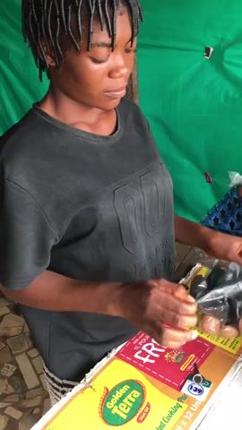 A young woman with braided hair, wearing a black t-shirt with embossed text that appears to read "DO" or "DD" followed by "SPORT", is standing behind a counter or table. She is situated in what looks like a market stall or small shop in Enyibichiri, Nigeria, with a vibrant green tarp serving as a backdrop.

On the table in front of her are various cardboard boxes and packaging materials. A prominent yellow box features the "Golden Terra" brand, and a red box clearly displays "FREE 1L POUCH" along with the text "Cooking Pan". The number "139" is also visible on one of the cardboard pieces, possibly indicating a price.

The woman is actively engaged in handling a black plastic bag with both hands, appearing to be in the process of opening it or preparing to package something. To her right, several tall stacks of colorful egg cartons (blue, purple, orange, and green) are neatly arranged, each filled with brown eggs. The woman's expression is calm and focused on her task.