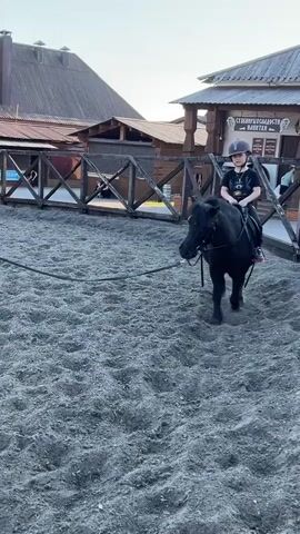 A boy wearing a helmet and black athletic clothes is riding a small black pony around a dirt arena. He is sitting on a saddle and holding the reins. A woman is walking alongside the pony, holding onto a lead rope attached to its bridle. The arena is surrounded by wooden fences and buildings with brown shingled roofs.  The ground is covered in gray sand.