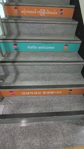 This media file shows a set of stairs decorated with welcome messages in different languages. The stairs are made of granite with black anti-slip strips on each step. There is a metal handrail on the left side of the stairs.

The visible text on the stairs is:
*   **Top stair (orange band):** "xin chao chao mung" (Vietnamese for "hello, welcome")
*   **Second stair from top (teal band):** "ສະບາຍດີ ຍິນດີຕ້ອນຮັບ" (Lao for "hello, welcome")
*   **Third stair from top (orange band):** "မင်္ဂလာပါ ကြိုဆိုပါတယ်" (Burmese for "hello, welcome")
*   **Fourth stair from top (teal band):** "Hello welcome" (English)
*   **Fifth stair from top (orange band):** "안녕하세요 환영합니다" (Korean for "hello, welcome")

Each band also features cartoon illustrations of children, suggesting a family-friendly or educational environment. The setting appears to be indoors, likely in a public building such as a school, community center, or office. The lighting suggests it is daytime. The overall impression is one of a welcoming and inclusive atmosphere, with efforts made to greet visitors in multiple languages. The location context provided is Asan-si, South Korea.