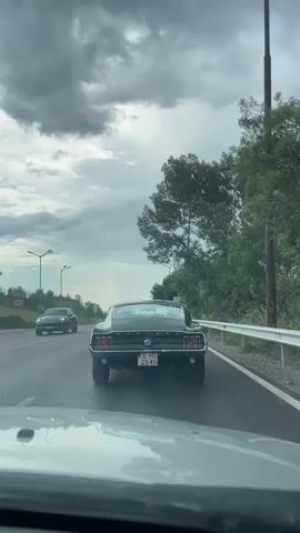 The video captures a view from a car driving on a road, following a vintage dark green Ford Mustang. The Mustang has a license plate with the text "E HT 2545". In the background, other cars are visible on the road, and to the right, there is a white guardrail and a dense line of trees. A blue sign is also visible, which reads "Драгичево" and "Dragichevo". The sky is overcast with dramatic, dark clouds, suggesting an impending storm or recent rain. The overall lighting indicates it is daytime, likely late afternoon or early evening. The scene appears to be a highway or a major road outside of a town or village, indicated by the presence of a bus stop sign and the town name sign. The activity is simply driving on a road, with the focus on the classic car leading the way. The mood is somewhat somber due to the dark sky, but also exciting due to the presence of the classic car.