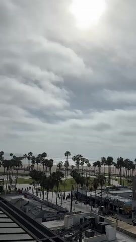 The video shows an aerial view of a beach boardwalk scene. In the foreground, there are rooftops of buildings, some with HVAC units and awnings. Beyond that, a street is visible with cars and a white van. Further back, a line of palm trees separates the street from a sandy beach, where the ocean meets the horizon. The sky is overcast with a bright sun peeking through the clouds. The overall impression is a bustling coastal urban environment.