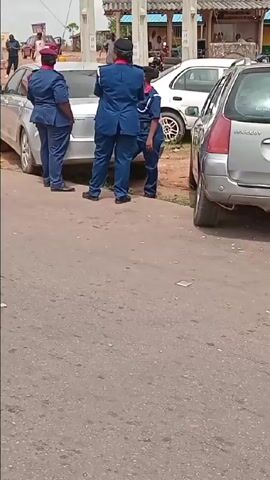 The video captures an outdoor scene near "THE NIGERIA POLICE FORCE NNPC OUTPOST KADUNA" in Ungwan Tanko, Nigeria. The time of day appears to be daytime, under an overcast sky.

The scene features numerous people, predominantly adults, gathered along a paved road. In the initial frames, several police officers in blue uniforms with red caps are seen standing near a silver car. As the video progresses, a growing crowd of men, women, and some children are visible. Many are sitting on concrete curbs along the roadside, while others stand in small groups, conversing or observing their surroundings. Some individuals are using mobile phones.

Various vehicles, including several cars (a silver sedan, a blue sedan, a red hatchback, and a black SUV), motorcycles, and a tricycle (Keke NAPEP), are present on the road or parked. One motorcycle is seen carrying multiple passengers.

People are dressed in a mix of casual and traditional Nigerian clothing. Scattered leaves are visible on the ground. A woman at 0:23 holds a sign, with "Take..." being faintly legible. The overall mood appears calm, suggesting a gathering or a period of waiting rather than an active event. Green trees and foliage are visible in the background, along with industrial structures of what appears to be the NNPC facility.