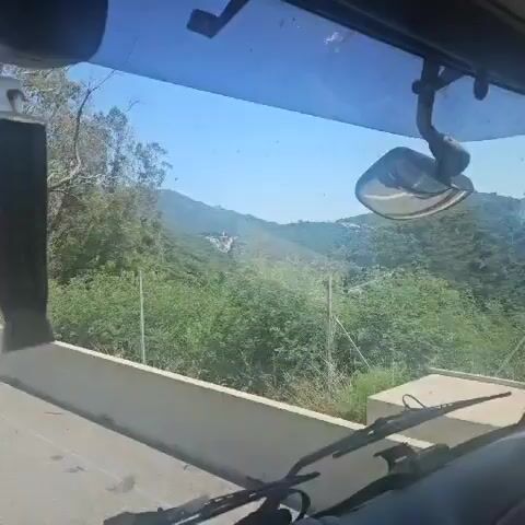 The video shows a landscape with a village nestled in a forest, viewed from inside a moving vehicle. The vegetation is primarily green trees and shrubbery. The village consists of several buildings with light-colored facades and red-tiled roofs. A tall, weathered stone tower rises above the buildings, and a white, monument-like structure stands next to the village. The backdrop is a hazy blue sky. The view is partially obstructed by the vehicle's window and the side mirror.  A concrete barrier and vertical metal posts are visible in the foreground.