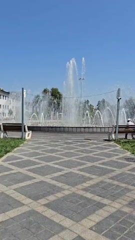 The video opens with a shot of a large fountain spraying water high into the air under a clear blue sky. There are some trees and buildings in the background. Two benches are visible in the foreground on either side of a paved walkway. A person is sitting on the bench to the right. The camera then pans to the left, showing a grassy area with several young trees. A few people are walking in the distance. The camera continues to pan, revealing more trees and buildings, including what appears to be a supermarket. The video ends with a shot of a single young tree standing in a grassy field.
