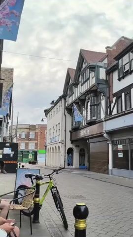 The video pans from left to right, showing a street scene with buildings on either side. A neon green bicycle is parked on the left side of the street, leaning against a metal post. A black and gold bollard is in the foreground. In the background, there are several shops, including "F HINDS" and "Clarks". A silver car is parked in front of the "Clarks" store. Further down the street, there is a shop called "mobile hut". The sky is cloudy.
