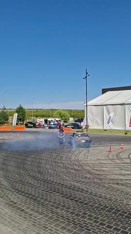 A person in a wheelchair is positioned in the center of a circular paved area surrounded by what appears to be a drift track. A black car is actively drifting around the person, creating a significant amount of smoke. Other cars, some of which appear to be drift cars, are parked in the background to the left of the frame. Spectators can be seen near these parked cars. A large white tent structure is visible on the right side of the frame. The sky is clear and blue. The scene suggests an automotive event or demonstration.