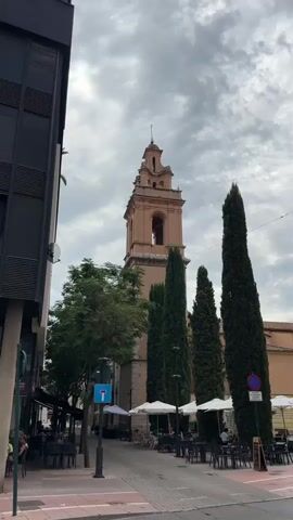 The video shows a tall, peach-colored bell tower with an arched opening and a cross at its peak, set against a cloudy sky. In front of the tower, there are several tall, slender green trees, and to the left, the side of a dark building. The ground level features an outdoor seating area with tables and chairs, some covered by white umbrellas, indicating a cafe or restaurant. A few people are seated at the tables. A blue street sign with a white arrow is visible on a lamppost.