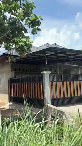 The video shows a moving camera perspective, likely from a vehicle, traveling along a dirt road beside a residential area. On the left, a fence with vertical orange and black slats encloses a grey, unfinished house with a dark tiled roof. The house has several rectangular windows and small rectangular vents near the roofline. Adjacent to the house, a grey concrete wall with small, evenly spaced rectangular openings runs along the property line. To the right of the road, there is dense green vegetation, including large-leafed plants resembling banana trees, and a tall tree with lush green foliage. The camera continues to move, revealing more of the greenery and passing by another section of a stone wall. Further along, the view shifts to show a white house with a beige trim and a dark tiled roof, also surrounded by greenery. The sky is blue with scattered white clouds. The overall impression is of a rural or semi-rural setting with residential properties interspersed with abundant vegetation.