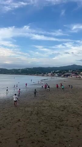 The video shows a beach scene with a group of people playing in the shallow water and on the wet sand. The sky is blue with scattered clouds. In the background, there is a coastline with hills covered in vegetation and buildings. The people, mostly children and some adults, are scattered across the beach. Some are running, some are standing, and some appear to be playing a game. The waves are small, and the water is calm. The sand is dark and wet, indicating that the tide is likely out or receding. The overall atmosphere is one of casual recreation and enjoyment of the beach.