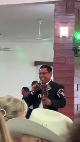 Here's a description of the video:

The video shows a man in a mariachi suit, holding a guitar and microphone, speaking at a gathering. He wears glasses and smiles as he addresses the crowd. 

The setting appears to be a banquet hall or large room with a low ceiling and ceiling fans.  A wooden pillar is visible, decorated with lighting equipment that includes green and amber lights. People sit at tables in the background. The foreground contains a partially visible audience including a woman with a hair clip and someone wearing a white cowboy hat.