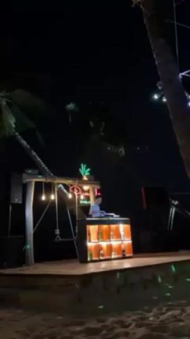 A DJ is playing music at an outdoor venue at night, likely a beach bar. The stage is wooden, lit with lights, and features a bar stocked with liquor bottles. Palm trees surround the area, contributing to a tropical atmosphere. The sand of the beach is visible in the foreground.