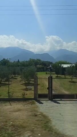 The video is a landscape shot taken from a moving vehicle, likely a car or bus, as indicated by the blurriness and the streaks of light reflecting on the window. The scene depicts a rural or semi-rural environment under a clear blue sky with some white clouds. In the foreground, there is dry, brown grass and a few bushes. Behind that, there is a field with trees, some of which appear to be fruit trees, planted in rows and fenced with simple posts. Further in the background, there are mountains. There are also some structures visible, including what appears to be a multi-story building on the left side and possibly a small open-air structure with a roof on the right. Power lines are visible across the sky. The sun is shining brightly, casting strong shadows and creating lens flares. The overall impression is of a sunny day in a mountainous region with some agricultural activity. There are no people clearly visible in the frame.