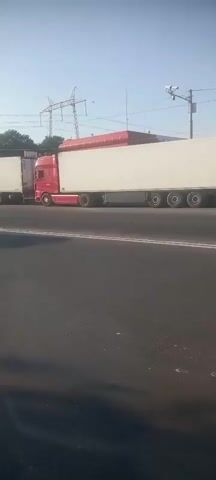 A silver sedan is seen driving on a highway. Behind it, a red truck with a long white trailer is also driving. The sky is clear and blue, and there are power lines in the background. The truck appears to be much larger than the car. The video is taken from a moving vehicle, likely another car on the highway, as evidenced by the motion blur and the perspective. The road surface is asphalt.