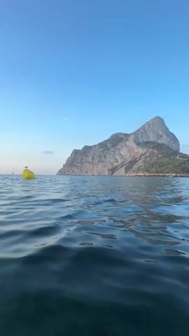 The video shows a low-angle shot of the ocean with a yellow buoy in the foreground. A seagull is perched on top of the buoy. In the background, a large, rocky mountain juts out of the water, and the sky is a clear blue with a few wispy clouds. The water is calm with small ripples. There are also two sailboats visible in the distance. The scene appears to be set at either sunrise or sunset due to the soft light.