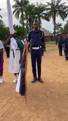 A medium shot shows people in uniforms standing at attention outdoors. The location appears to be a yard with red dirt. There are palm trees in the background. In the center is a young man in a dark blue uniform with yellow epaulets and a dark blue hat with a yellow brim. He holds a tall flag with a dark blue stripe, a white stripe, and a golden fringe. To his left, another person wears a white tunic, a green cape, and a golden crown. They hold a tall white flag. Other people in similar dark blue uniforms stand in formation in the background, along with people in other kinds of uniforms. The sound of a drum can be heard.