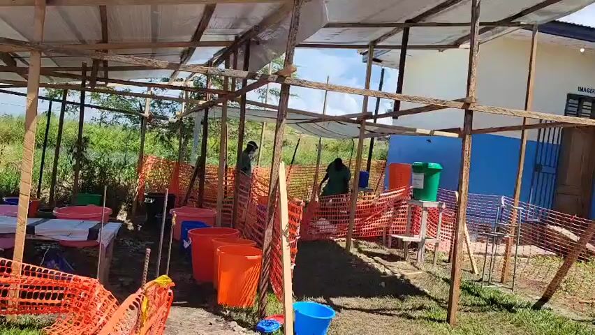 The image depicts an outdoor, semi-covered facility, likely a hygiene or processing station, set up in Tchere, Democratic Republic of the Congo. A wooden frame structure with a translucent plastic sheet roof provides partial shelter. Orange safety netting is extensively used to create barriers and delineate areas within the facility.

Two individuals, possibly men, wearing dark green uniforms or work clothes, are present. One is standing near a hand-washing station on the right, which consists of a green barrel with a tap placed on a stand next to an orange bucket. The other person is further back, moving within the netted area.

Numerous plastic buckets in various colors (red, orange, blue, green) are scattered throughout the space, some seemingly in use. On the far left, a white table or counter holds several items. In the background, a blue building with a white upper section and a brown door is visible. Above the door, the word "IMAGERIE" is clearly written, indicating it is likely an imaging or radiology department, suggesting the overall facility might be part of a medical or health center.

The setting is outdoors, with bright natural light suggesting it is daytime and sunny. Lush green foliage is visible beyond the structure. The overall arrangement points to an organized, temporary, or semi-permanent facility focused on health or sanitation.