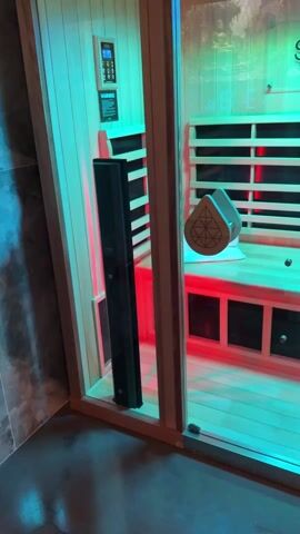 The camera pans across a room focusing on a wooden-framed glass-walled sauna. Inside the sauna, the wood is lit by color-changing LEDs cycling through red, green, and blue. There is a small control panel on the side wall. The sauna has wooden benches, and a decorative wooden handle is on the glass door. The camera then shifts to show a grey tile shower stall adjacent to the sauna, with a lit mirror on the wall and a glass door. The floor is also tiled.