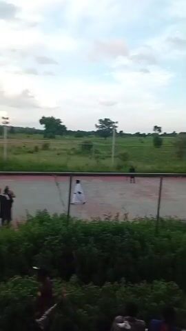 The video shows an outdoor sports court, possibly a tennis court, with a net across the middle. There are people on the court, some appear to be playing a game, while others are standing around. A crowd of spectators is visible in the foreground, seated on bleachers or a raised area. The environment appears to be a field or open space, with trees and a cloudy sky in the background. There are lights for the court, and a fence surrounds it. The overall scene suggests a sporting event taking place.