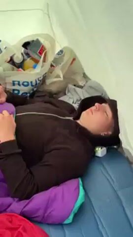 Here is a concise description of the video:

The video starts with a shot inside a tent where a person is lying asleep in a sleeping bag. The tent is cluttered with bags and other camping essentials. The camera then moves to show the outside of the tent revealing a bustling campsite filled with tents of various colors and sizes. Two people are sitting outside the tent in camping chairs, enjoying the scene. One person wears sunglasses, while the other is focused on a mobile phone. The sky is blue with scattered clouds, and colorful bunting decorates the area. The scene captures a relaxed atmosphere at a festival or camping event.