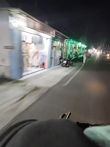 The video shows a nighttime scene from the perspective of someone on a motorcycle. The motorcycle is moving along a street lined with buildings. The buildings have awnings adorned with green decorative lights and banners. Several motorcycles are parked along the sidewalk in front of the buildings. A person in light-colored clothing is walking on the sidewalk. The street is paved, and there are streetlights in the distance. The overall impression is a moderately lit urban or suburban environment at night.