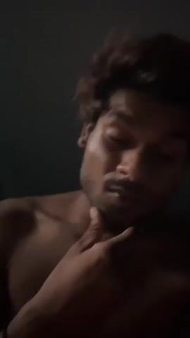 A close-up, low-light video shows a person with dark hair and stubble. The person appears to be male and is shirtless. The person's right hand is raised to their head and neck area, running through their hair. The background is dark and indistinct.