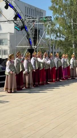 A group of twelve women in traditional folk attire, consisting of long skirts, vests, and head coverings, stand in a line outdoors. They appear to be singing, as some are holding their hands near their mouths. Behind them, a stage structure with lighting and speakers is visible, along with a modern building and trees under a clear blue sky. The ground is paved with light-colored square tiles.