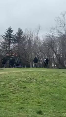 The video shows a group of people on what appear to be electric bikes or scooters gathered on a grassy hill in a park-like setting. The weather is overcast. Some of the people are standing by their vehicles, while others are seated. In the distance, there are trees and buildings. The camera pans to the right, showing more of the park, including other people walking around. A body of water, possibly a lake or bay, is visible in the background.