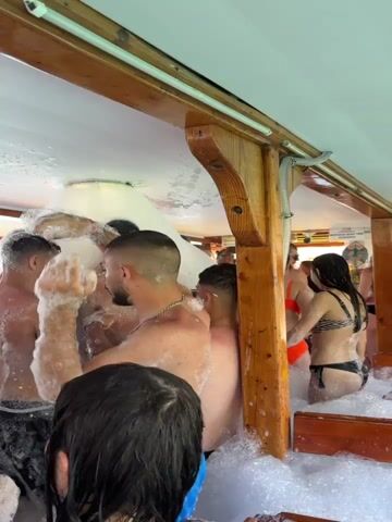 The video captures a lively foam party taking place inside what appears to be a party boat in Menderes, Turkey. Numerous young adults, both male and female, are seen covered in white foam from head to toe.

The boat's interior features a wooden ceiling and support beams. A foam machine, mounted to the ceiling, is actively dispensing a large volume of foam, which fills the space, creating a thick, sudsy environment. Most of the participants are shirtless or wearing swimwear, with their hair and bodies saturated in foam. They are standing closely together, moving to the upbeat music playing in the background.

People appear to be enjoying the event, smiling and interacting. One man in the foreground is seen wiping foam from his face, while others are swaying and dancing. In the background, a woman in a striped bikini top and dark bottoms is visible. The lighting is bright, suggesting it is daytime. On the right side, a sign with partially visible text, possibly related to boat tours, can be seen. The overall scene depicts a fun and energetic party atmosphere on the water.