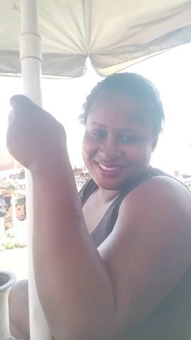 A woman is standing under an umbrella and smiling into the camera. She is wearing a dark tank top. She is holding the umbrella pole.  Other people are visible in the background, as well as items for sale.
