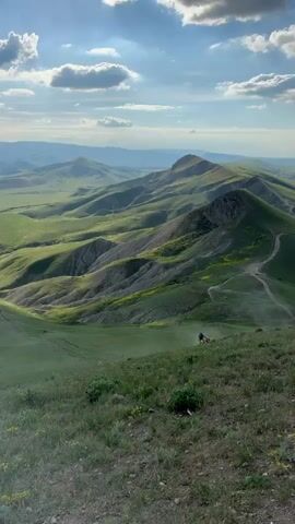 The video shows a vast hilly landscape under a partly cloudy sky. The hills are covered in green grass with patches of yellow flowers, and there are visible dirt paths winding across the terrain. In the distance, mountains can be seen on the horizon. Two motorcycles are visible in the middle of the frame, riding along the grassy hills. The viewpoint is from an elevated position, overlooking the scene.