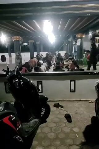 The video shows a group of people, mostly young males, gathered in an indoor space that appears to be a place of worship. They are dressed in casual clothing and some are wearing skullcaps. Many individuals are kneeling or bowing in prayer, facing away from the camera. The environment is dimly lit, with overhead lights providing illumination. In the foreground, two motorcycles are parked, and several pairs of sandals are scattered on the floor. The background shows columns and a wall with rectangular openings. Some individuals are standing or walking in the background. The overall impression is of a religious gathering in progress.