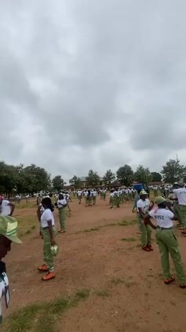 The video depicts an outdoor scene with a large group of young adults in matching green and white uniforms. They are spread across a dirt field with some grassy areas and trees in the background. In the distance, there is a covered structure with a stage or platform, adorned with Nigerian flags. Many individuals are wearing white t-shirts with "NYSC" visible on the back, and green caps. Some are standing, while others appear to be engaged in activities like dancing or marching. The sky is overcast with clouds. The camera is positioned at various angles, often focusing on different individuals within the group.