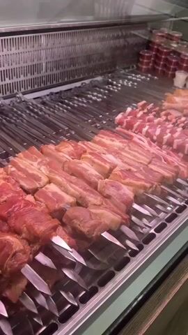 A close-up, angled shot shows rows of raw meat on skewers arranged on metal racks, likely in a display case. The meat appears to be pork belly and other cuts, cut into uniform pieces and threaded onto long metal skewers. In the background, smaller containers of sauces or dips are visible, stacked neatly. The lighting suggests a well-lit commercial environment, possibly a butcher shop or restaurant kitchen. The overall impression is one of food preparation and presentation.