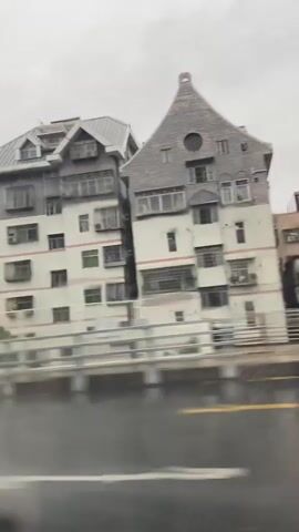 The video captures a rainy day from the perspective of someone inside a vehicle. The scene is characterized by a grey, overcast sky and wet roads, indicated by reflections and splashes from passing cars. Tall buildings and apartment complexes dominate the cityscape in the background, suggesting an urban environment. A dark SUV with a spare tire mounted on the back is seen driving on the road. There are also streetlights and traffic signs visible. The overall atmosphere is that of a typical rainy day commute in a city.